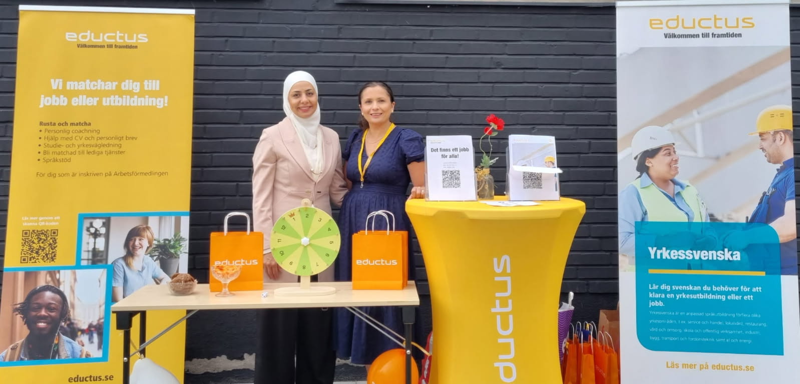 Eductus medarbetare deltagar i mässan på Hela Kronan gungar i Trollhättan. De står i sin monter med ett bord framför sig som är fyllt med profilprodukter och information om Eductus. På sidorna står två roll-ups som har information om Rusta och matcha samt yrkessvenska
