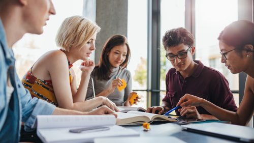 En engagerad grupp studenter sitter runt ett bord vid ett ljust fönster och studerar tillsammans med öppna böcker. Här ser vi värdet av samarbete när du läser Svenska med yrkesinriktning. Att studera SMY gemensamt förbereder dig för arbetslivet.